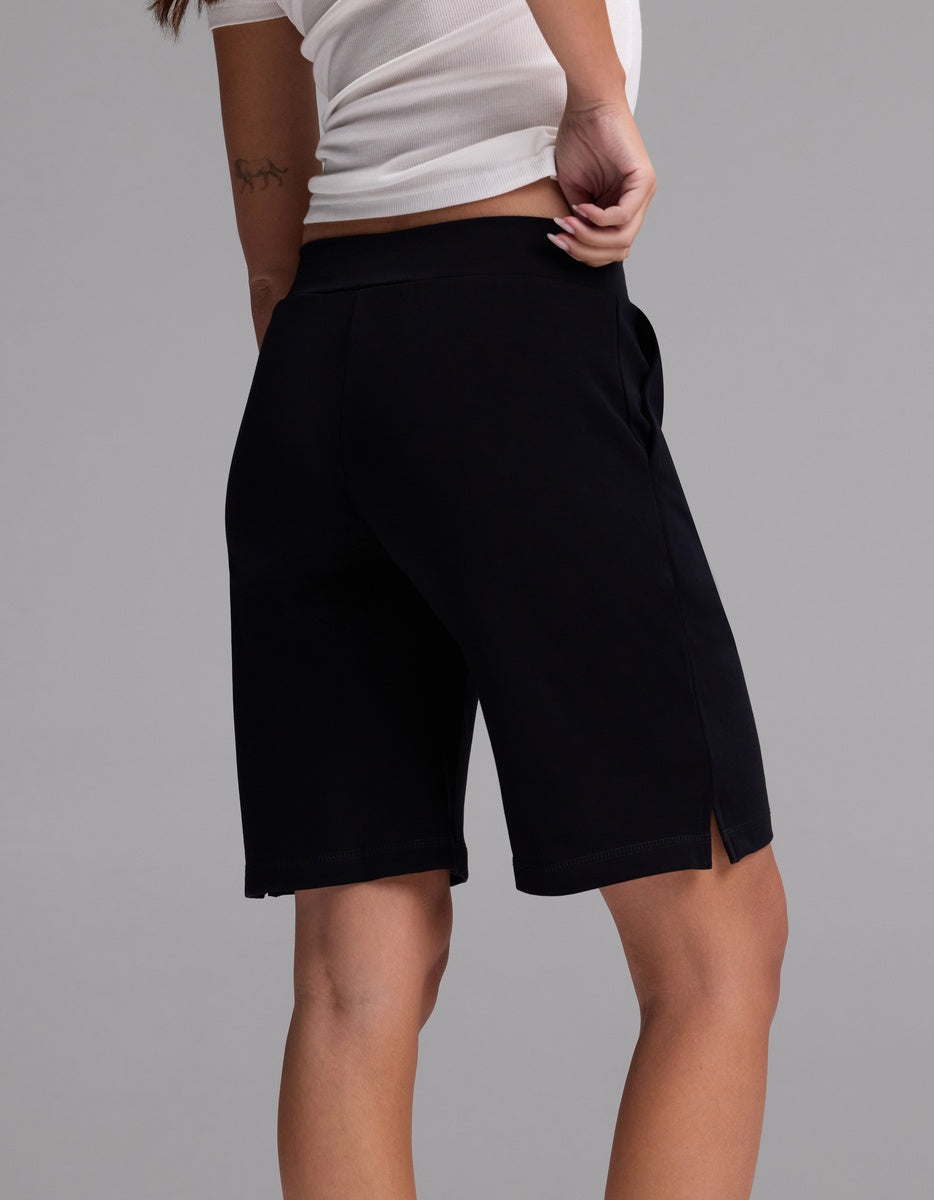 A person wearing a white shirt and Cozy Earth Women’s Brushed Bamboo Bermuda Shorts in black stands with one hand on their hip, facing away from the camera against a plain gray background.|Color:Black