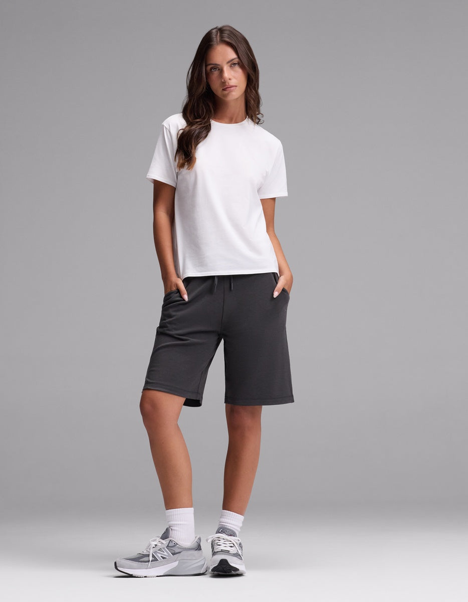 A woman stands against a gray background wearing Cozy Earth’s Women’s Brushed Bamboo Bermuda Shorts in dark gray, paired with a white t-shirt, white socks, and gray sneakers. Her hands are in her pockets as she looks slightly to the side.