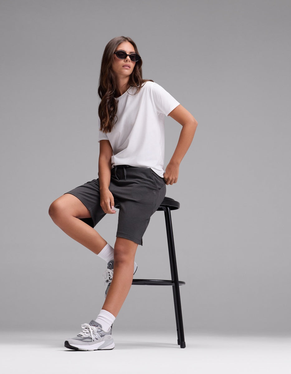 A person wearing sunglasses, a white t-shirt, white socks, gray sneakers, and Cozy Earth’s Women’s Brushed Bamboo Bermuda Shorts in dark gray sits casually on a tall black stool against a plain gray background.