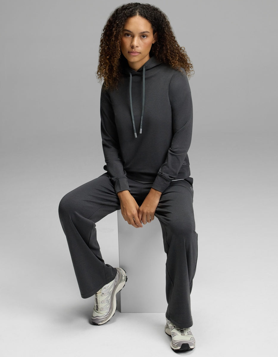 A woman with curly hair wears the Cozy Earth Women’s Brushed Bamboo Hoodie paired with matching pants, sitting on a white block and looking at the camera. She completes the look with white sneakers, set against a plain light gray background.