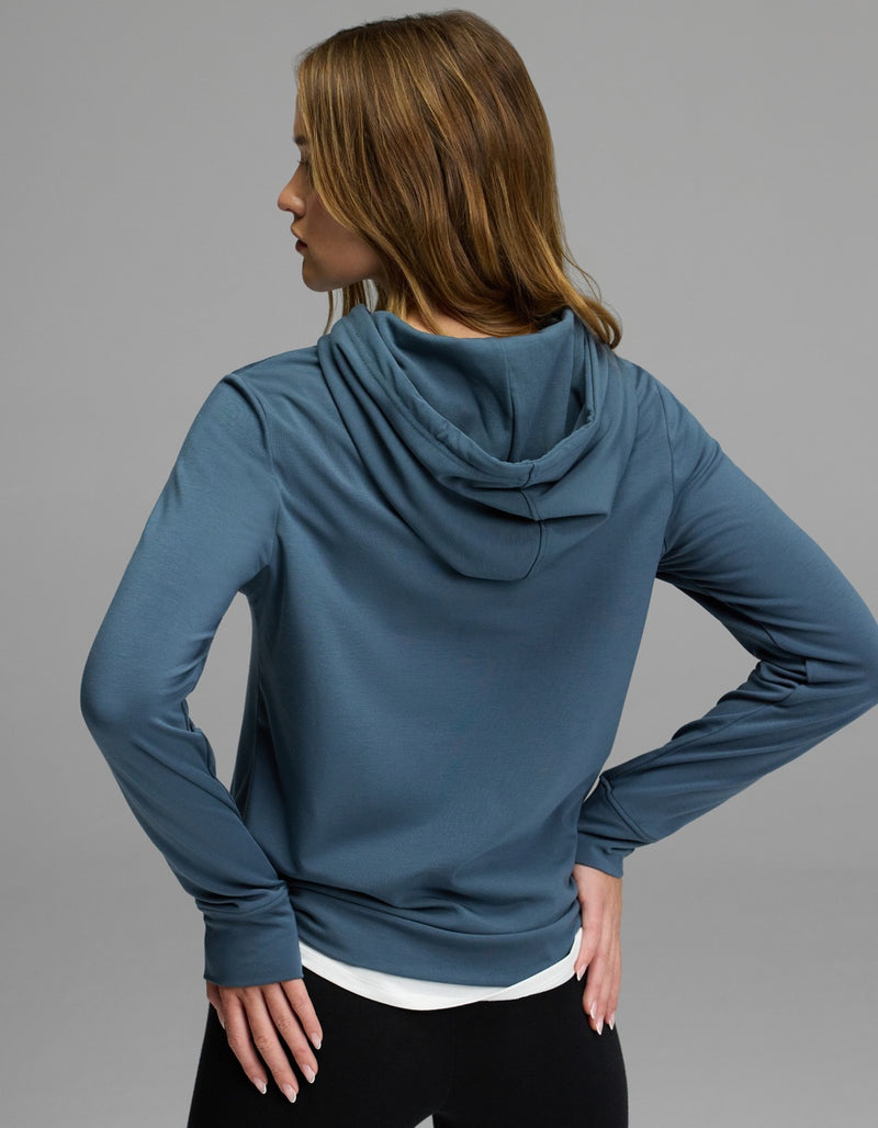 A person with long, light brown hair is seen from behind, wearing the Cozy Earth Women’s Brushed Bamboo Hoodie in blue and black pants, with one hand on their hip.
