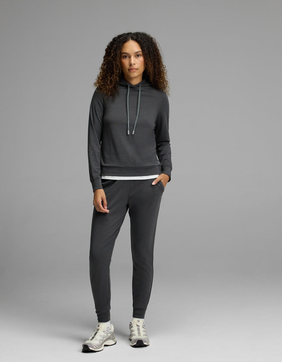 A woman with curly hair models the Cozy Earth Women’s Brushed Bamboo Hoodie & Jogger Set in a dark shade, paired with sneakers, standing against a plain gray background with a neutral expression.