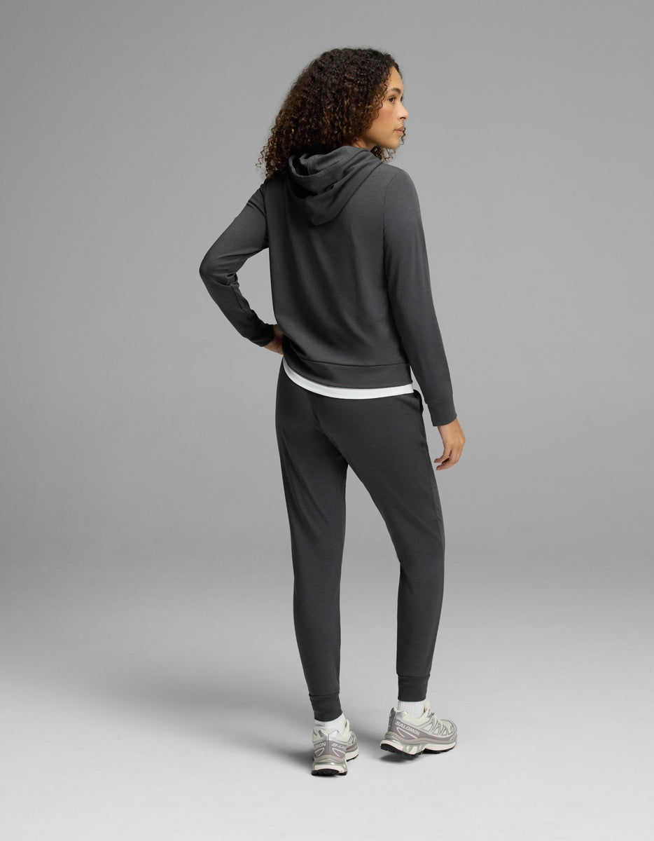 A woman with curly hair, dressed in the Cozy Earth Women’s Brushed Bamboo Hoodie & Jogger Set in a dark color, stands with one hand on her hip and faces away from the camera against a plain gray background.