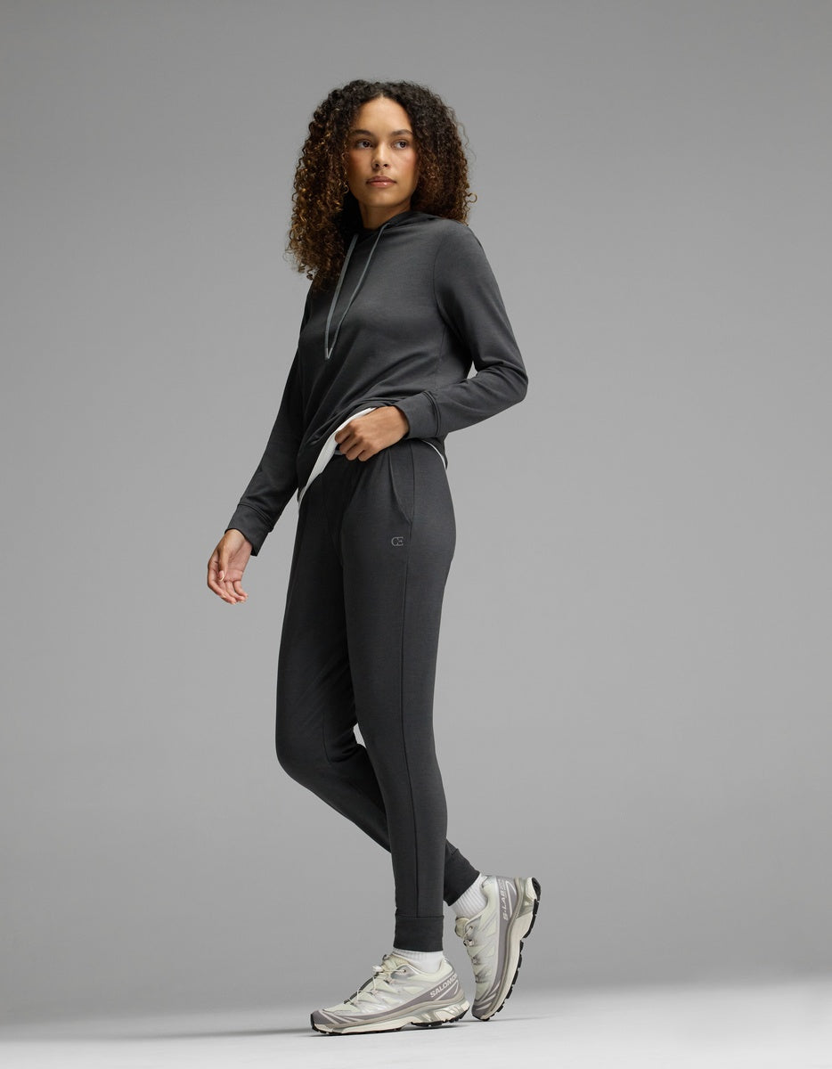 Wearing the Cozy Earth Women’s Brushed Bamboo Hoodie & Jogger Set in dark gray, a woman with curly hair pairs her outfit with white sneakers, standing against a plain gray background and gazing slightly to the side.