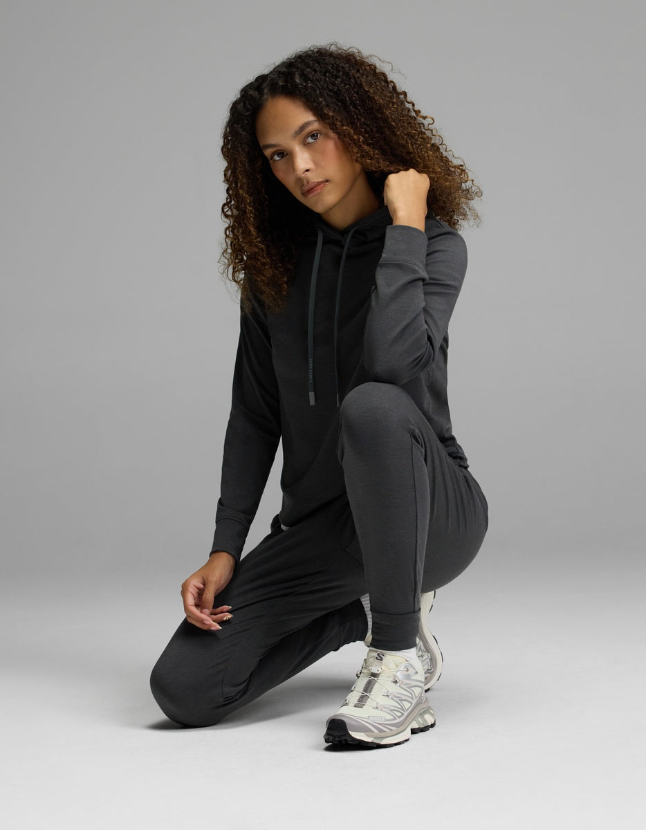 A woman with curly hair kneels on one knee against a plain gray background, wearing the Cozy Earth Women’s Brushed Bamboo Hoodie & Jogger Set in a dark color. She rests one hand on her chin and looks at the camera, completing her look with white sneakers.