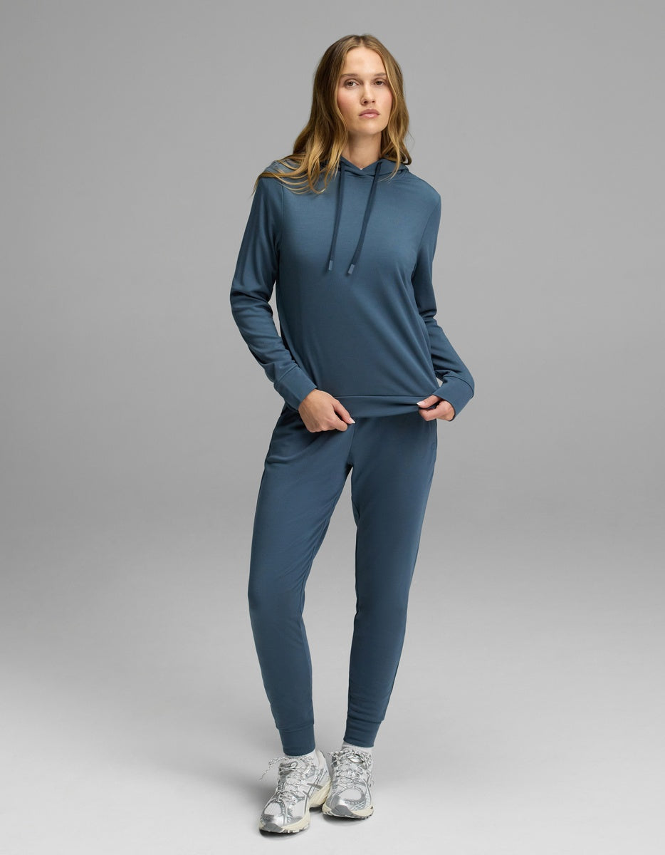 A woman with long blonde hair stands on a gray backdrop wearing the Cozy Earth Women’s Brushed Bamboo Hoodie & Jogger Set in blue, paired with white running shoes. She is holding part of her sweatshirt.