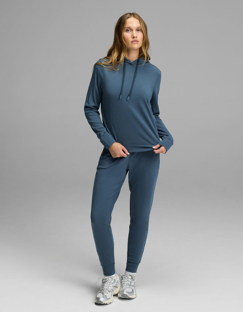 A woman with long blonde hair stands on a gray backdrop wearing the Cozy Earth Women’s Brushed Bamboo Hoodie & Jogger Set in blue, paired with white running shoes. She is holding part of her sweatshirt.