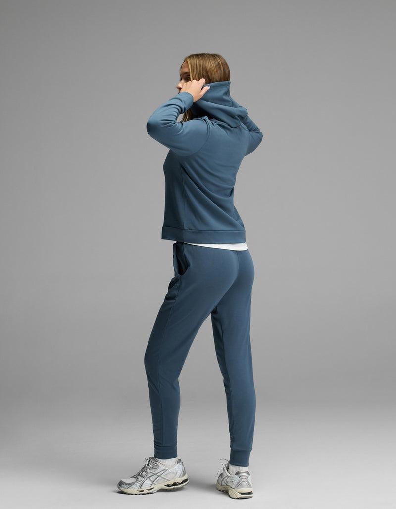A woman wearing the Cozy Earth Women’s Brushed Bamboo Hoodie & Jogger Set stands on a gray background, facing away as she pulls up the hood over her head.