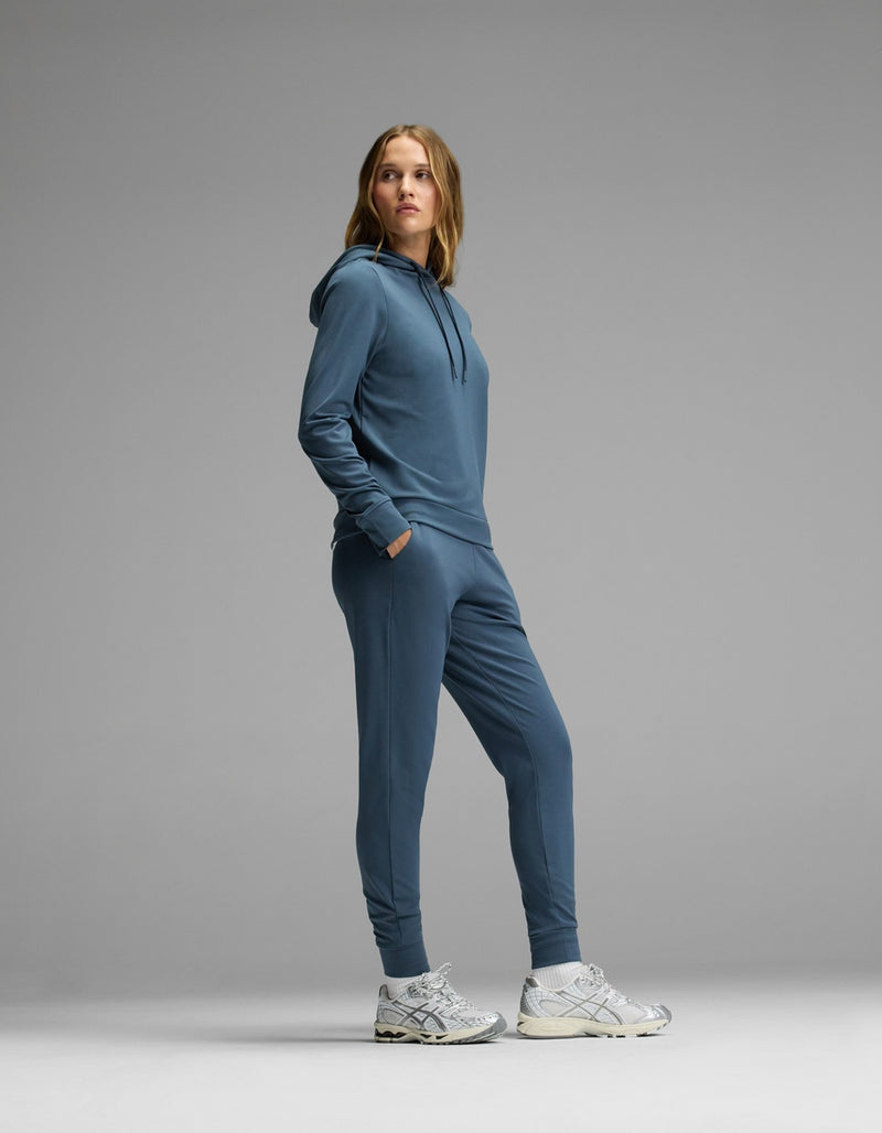 A woman stands sideways with her hands in the pockets of the Cozy Earth Women’s Brushed Bamboo Hoodie & Jogger Set, paired with gray sneakers, against a plain gray background.
