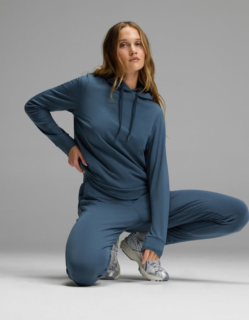 A woman showcases the Cozy Earth Women’s Brushed Bamboo Hoodie & Jogger Set in blue, kneeling on one knee with a hand on her hip against a plain gray background. She pairs the outfit with silver athletic shoes, and her long wavy hair cascades over her shoulders.