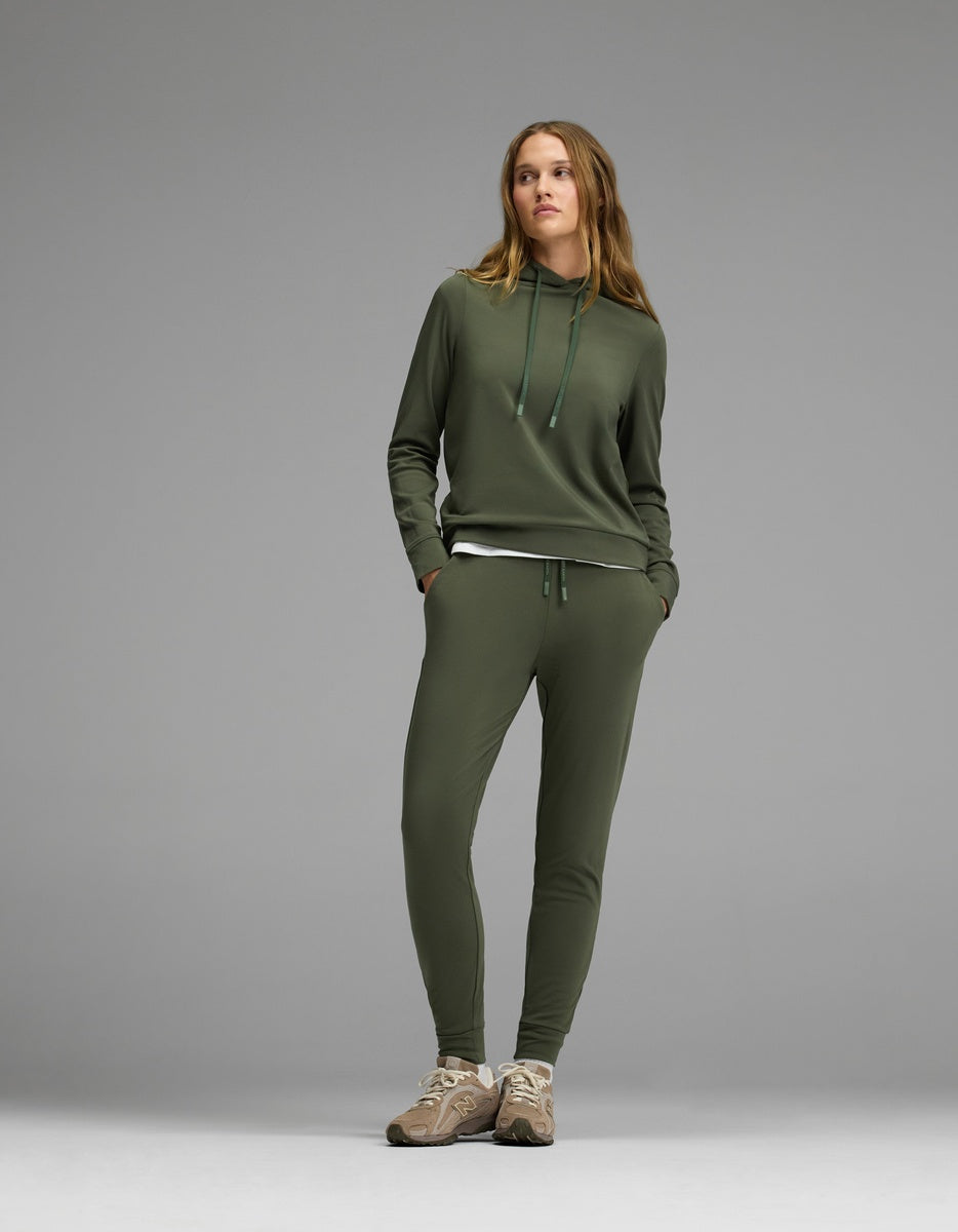A woman stands against a gray background wearing the Cozy Earth Women’s Brushed Bamboo Hoodie & Jogger Set in olive green, paired with beige sneakers. Her hands are in her pockets as she looks slightly to the side.