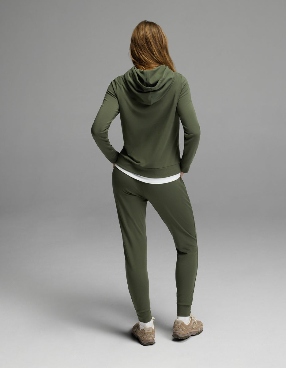 A woman with long brown hair, wearing the Cozy Earth Women’s Brushed Bamboo Hoodie & Jogger Set in olive green, stands facing away from the camera against a plain gray background.