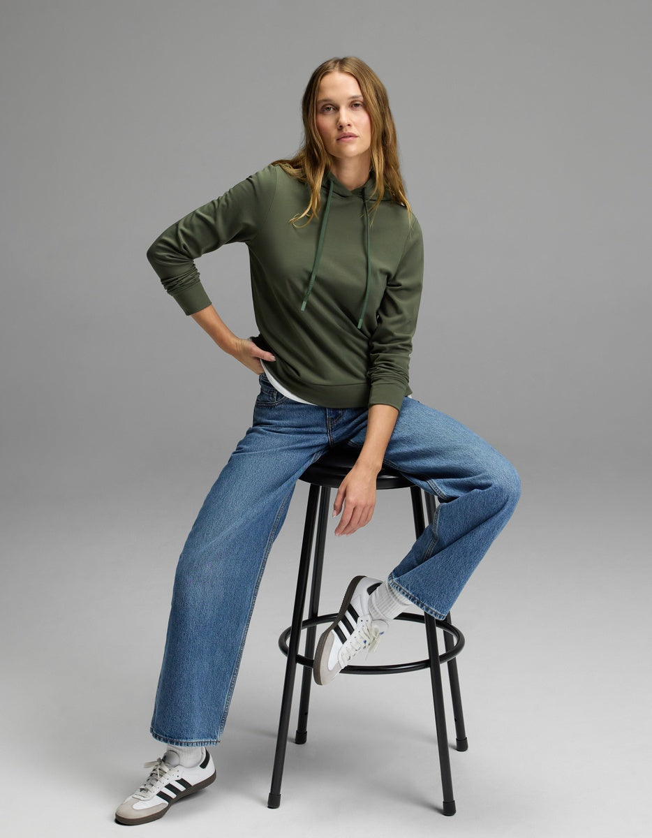 A person wearing the Cozy Earth Women’s Brushed Bamboo Hoodie, blue jeans, and white Adidas sneakers sits on a black stool against a plain gray background.