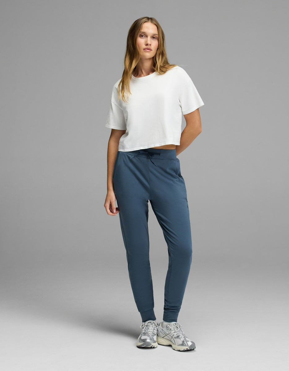 A woman stands facing forward against a plain gray background, wearing Cozy Earth’s Women’s Brushed Bamboo Jogger in blue, paired with a white cropped t-shirt and gray sneakers.