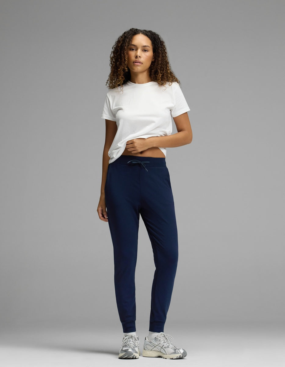 A woman stands against a plain gray background, wearing Cozy Earth’s Women’s Brushed Bamboo Jogger in dark blue, paired with a white t-shirt and gray sneakers. Her curly hair is down and she has one hand in her pocket.