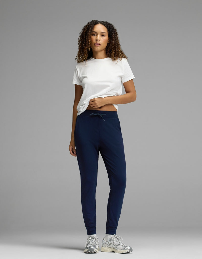 A woman stands against a plain gray background, wearing Cozy Earth’s Women’s Brushed Bamboo Jogger in dark blue, paired with a white t-shirt and gray sneakers. Her curly hair is down and she has one hand in her pocket.