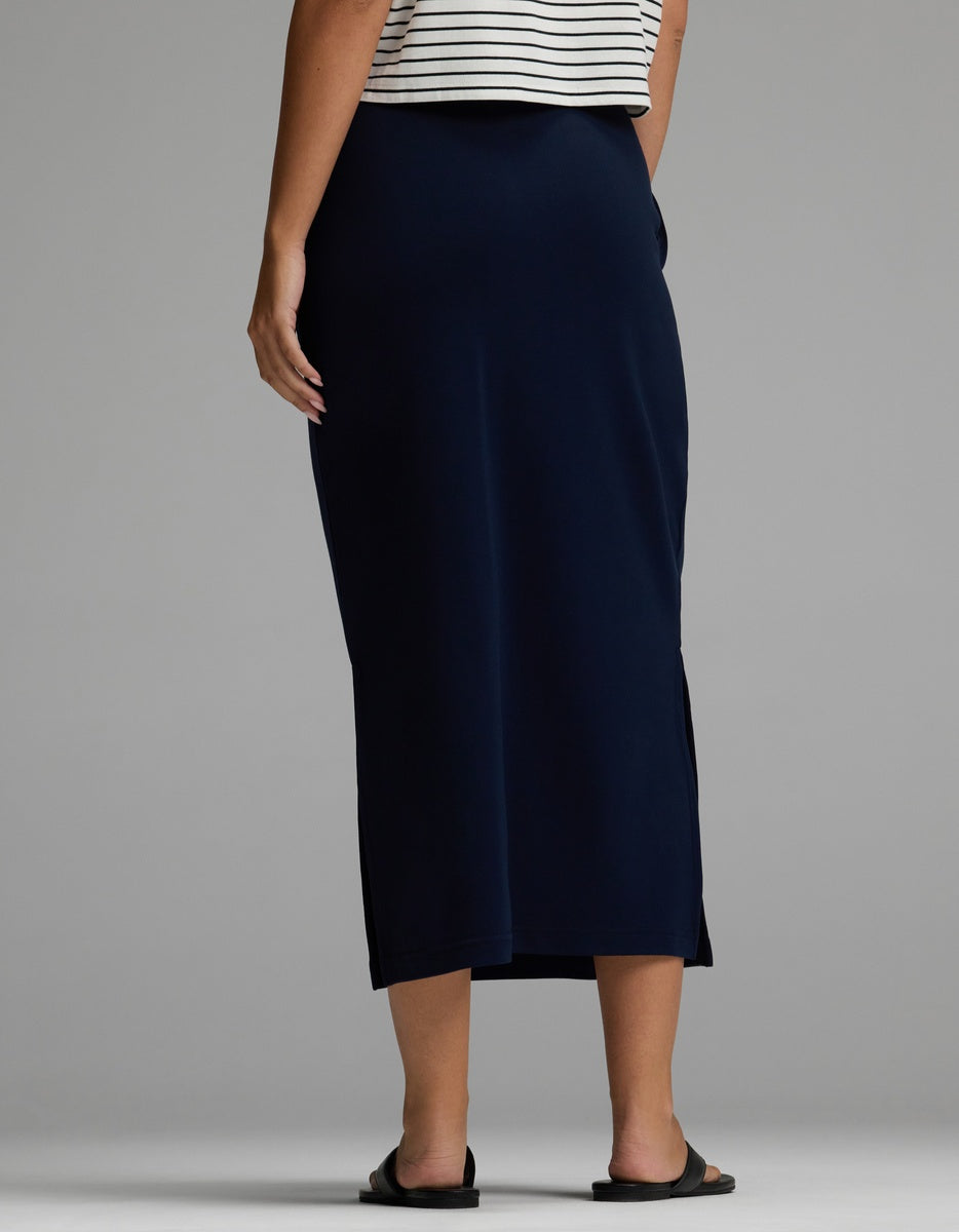 A person is shown from the back against a plain gray background, wearing the Cozy Earth Women’s Brushed Bamboo Midi Skirt in navy blue, paired with a striped top and black sandals.|Color:Navy