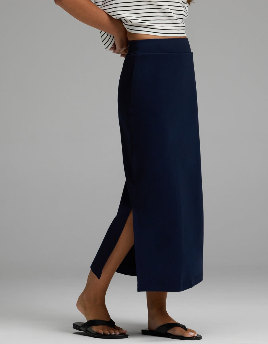 A person is wearing the Cozy Earth Women’s Brushed Bamboo Midi Skirt in navy blue with a side slit, paired with black slide sandals and a cropped striped top, standing against a plain gray background.|Color:Navy