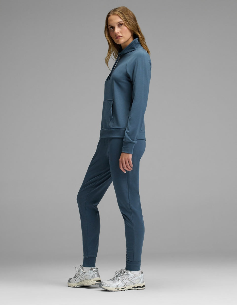 A woman stands sideways against a gray background, wearing Cozy Earth’s Women’s Brushed Bamboo Mock Neck with white sneakers. She looks at the camera with a neutral expression.