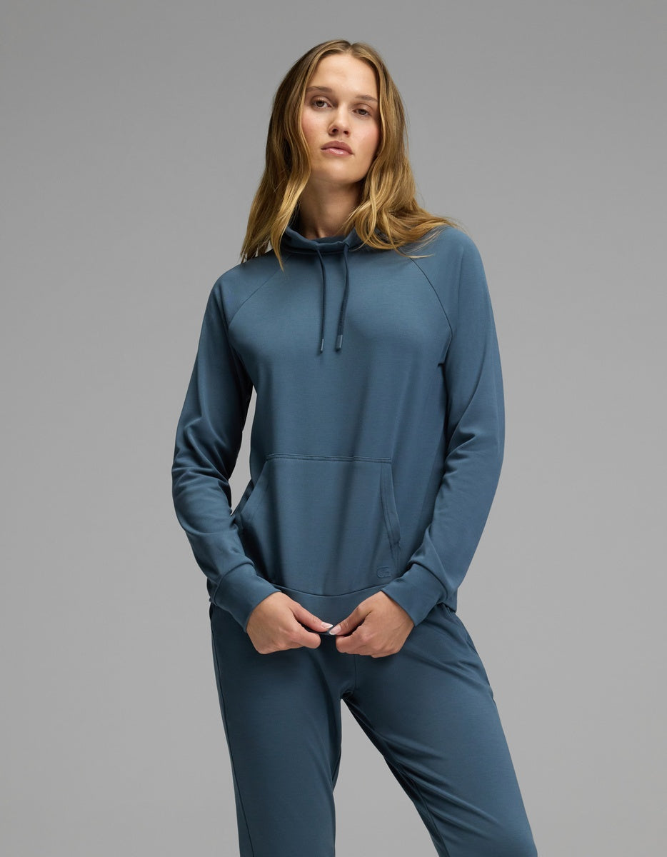 A woman models the Cozy Earth Women’s Brushed Bamboo Mock Neck in teal, standing confidently with her hands in the pockets against a plain gray background.