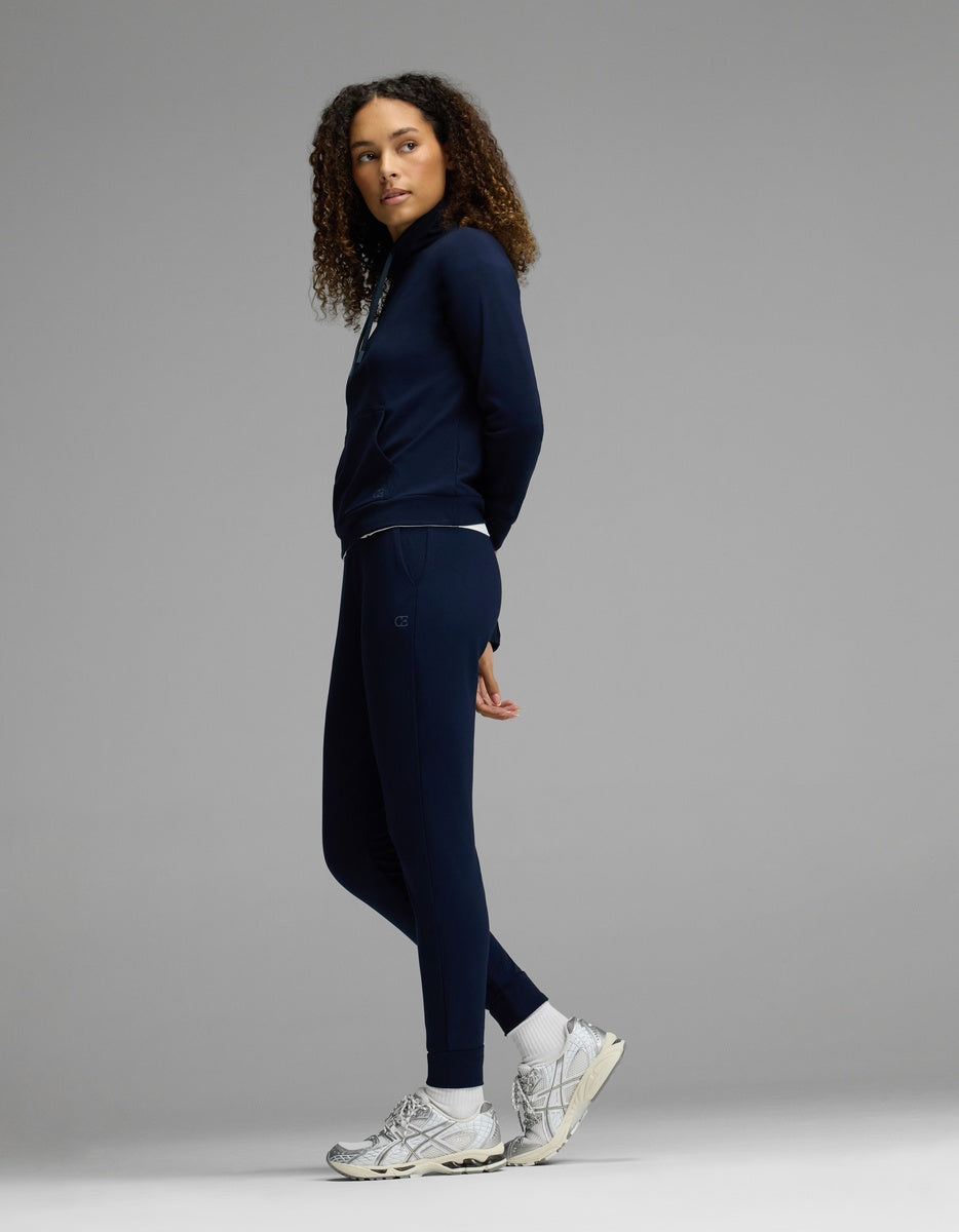 A person with curly hair stands sideways against a plain gray background, wearing Cozy Earth’s Women’s Brushed Bamboo Mock Neck & Jogger Set in navy, paired with white sneakers.|Color:Navy