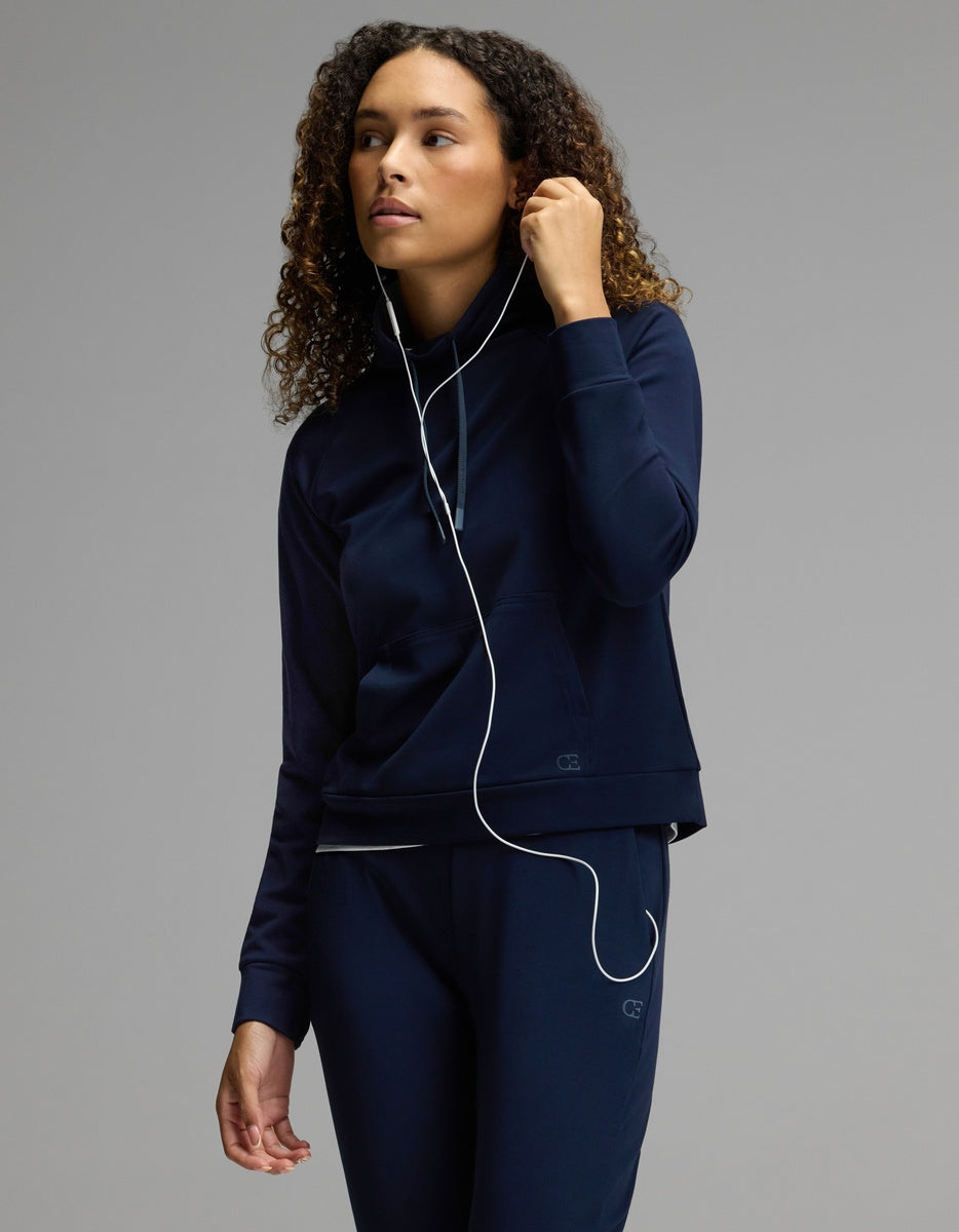 A woman with curly hair, dressed in the Cozy Earth Women’s Brushed Bamboo Mock Neck & Jogger Set in navy, listens to wired earphones—holding one earbud and gazing to the side against a plain gray background.