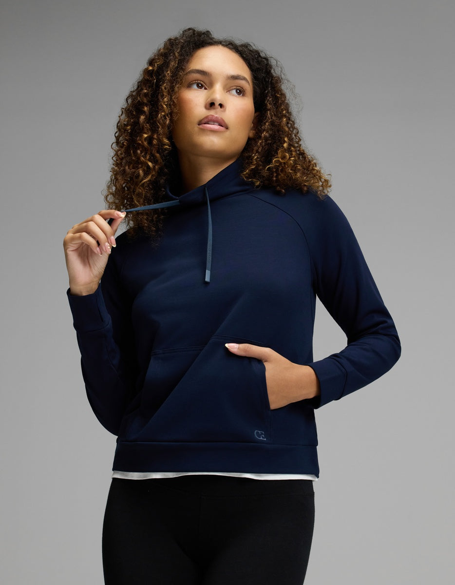 A woman with curly hair wears the Cozy Earth Women’s Brushed Bamboo Mock Neck in navy, paired with black leggings. She gazes upward and slightly to the side, holding a drawstring with one hand while her other hand rests in the front pocket, set against a neutral gray background.|Color:Navy