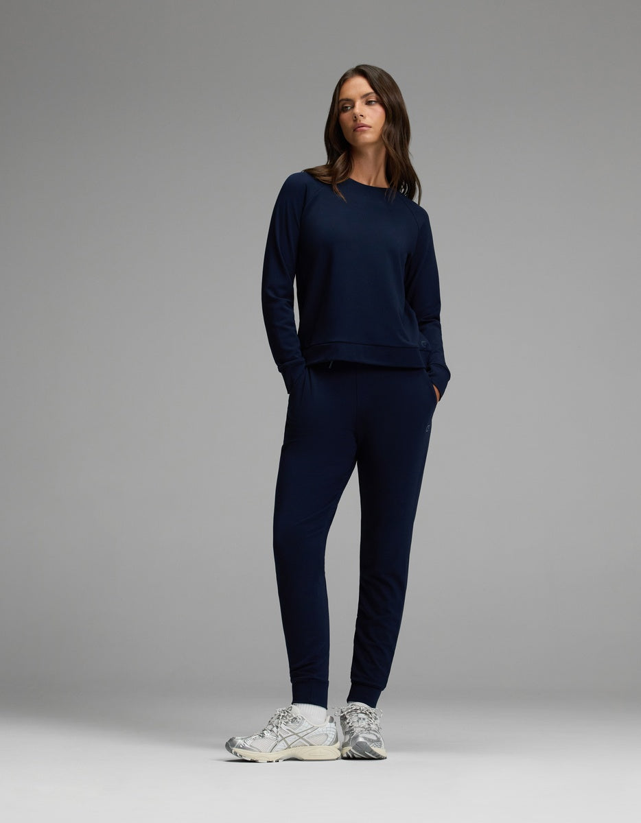A woman models the Cozy Earth Women’s Brushed Bamboo Jogger Set in Navy, featuring a long-sleeve top and matching jogger pants with hands in the pockets, paired with white sneakers against a plain gray background.|Color:Navy