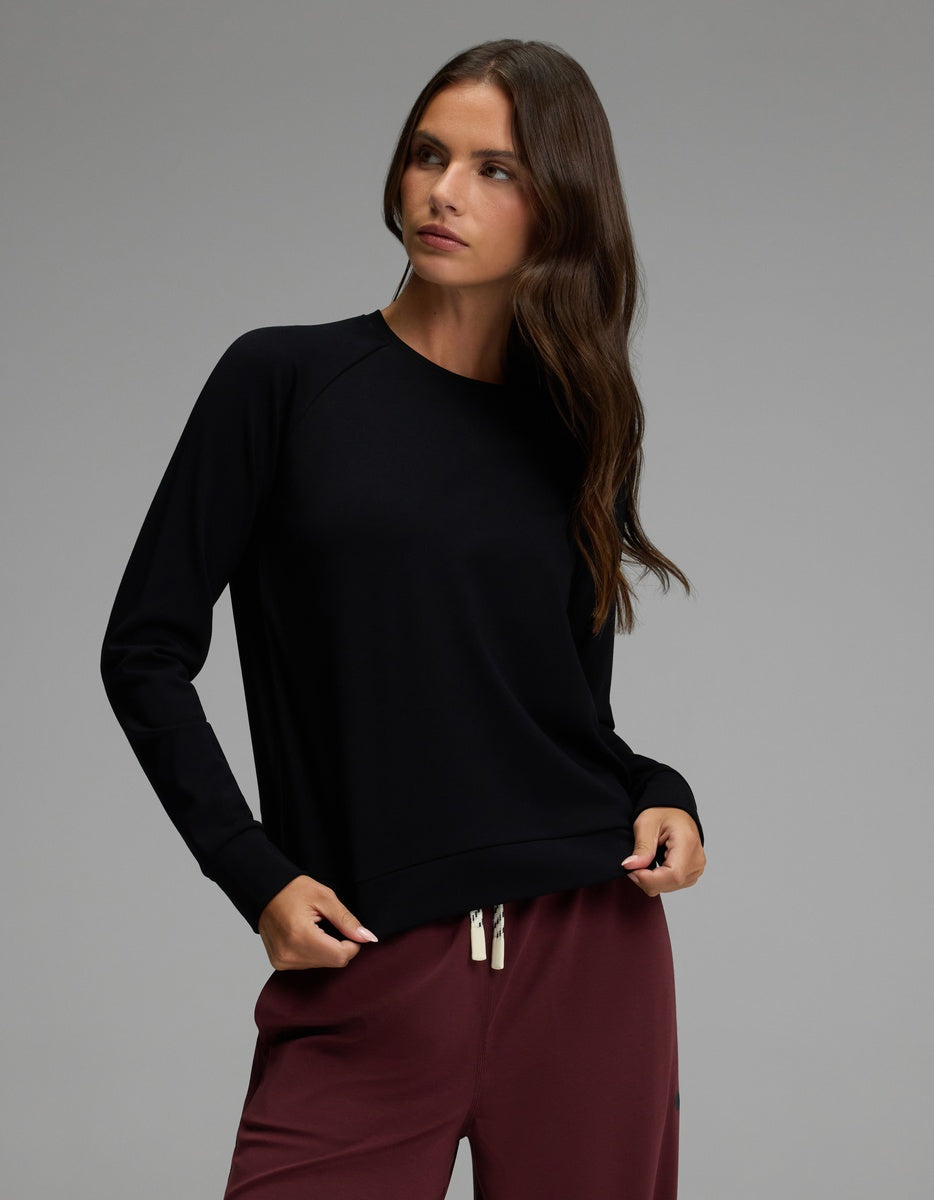 A woman with long brown hair wears the Cozy Earth Women’s Brushed Bamboo Pullover Crew in black and maroon drawstring pants, standing against a plain grey background and looking to her right with a neutral expression.|Color:Black