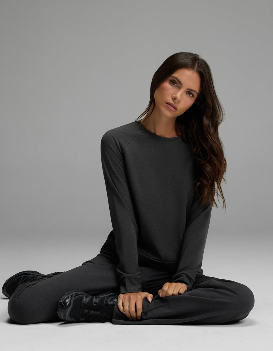 A woman with long brown hair sits cross-legged on the floor, wearing the Cozy Earth Women’s Brushed Bamboo Pullover Crew and matching pants, looking slightly to the side against a plain gray background.|Color:Charcoal