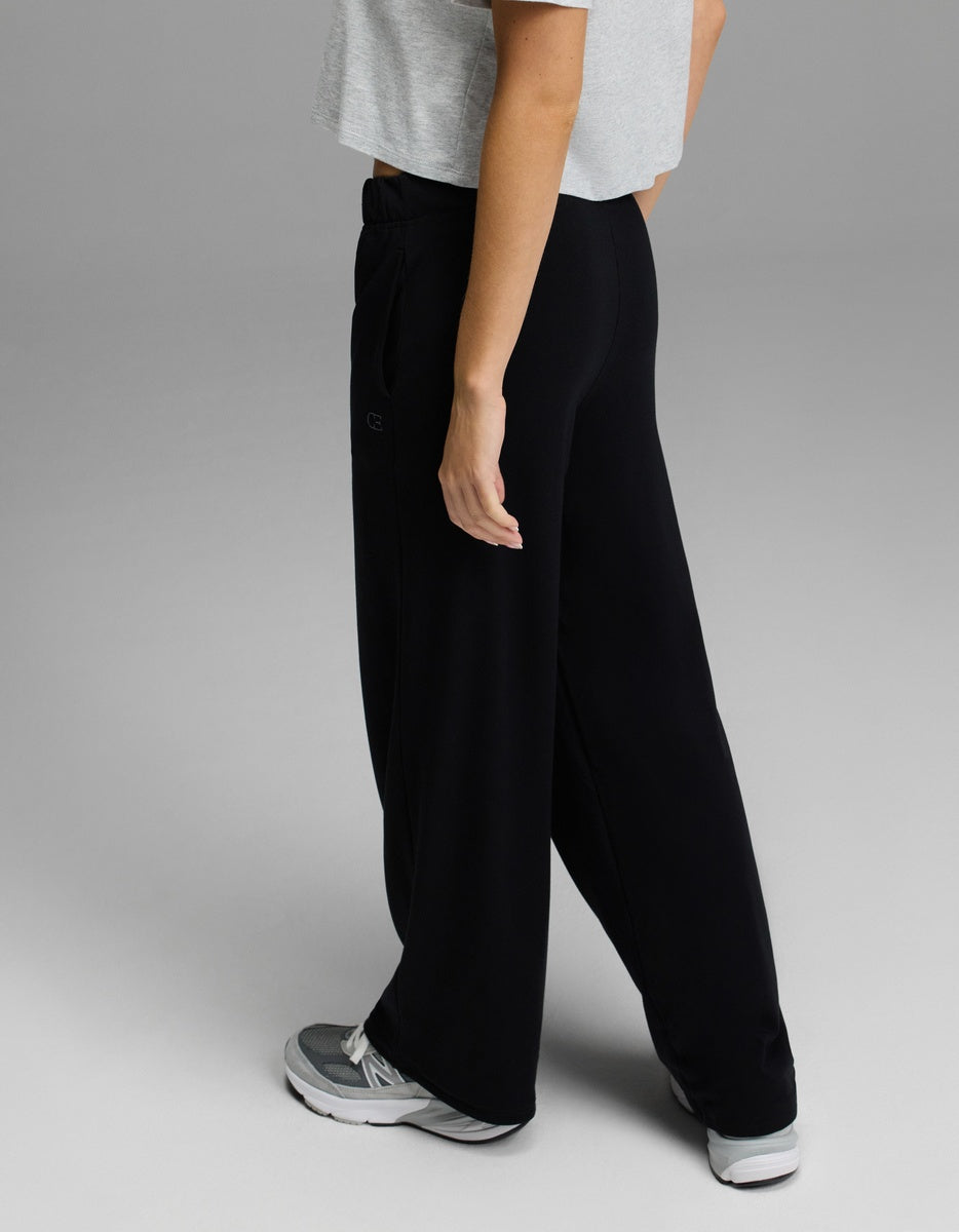 A person is shown from the side, cropped from the chest up, standing in a relaxed pose against a plain gray background while wearing loose black Cozy Earth Women’s Brushed Bamboo Wide Leg Pull-On Pants, a gray short-sleeve top, and gray sneakers.