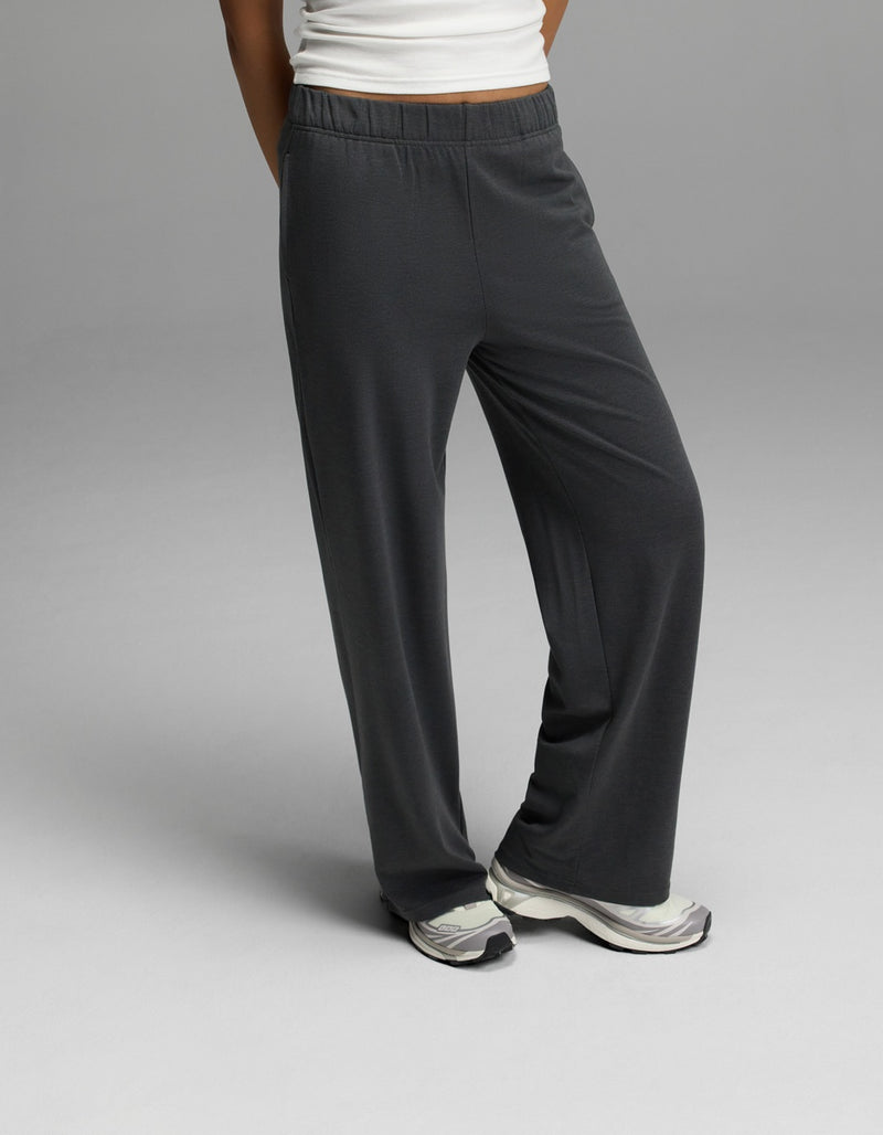 A woman wears Cozy Earth’s Women’s Brushed Bamboo Wide Leg Pull-On Pant in Charcoal, paired with white sneakers and a white top, standing with one leg crossed in front of the other against a plain gray background.
