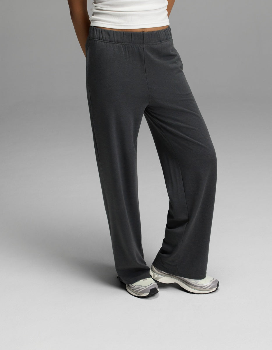 A woman wears Cozy Earth’s Women’s Brushed Bamboo Wide Leg Pull-On Pant in Charcoal, paired with white sneakers and a white top, standing with one leg crossed in front of the other against a plain gray background.|Color:Charcoal