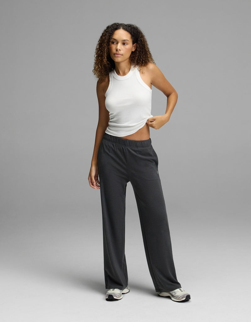 A woman with curly hair stands against a plain gray background, wearing a white sleeveless top and the Cozy Earth Women’s Brushed Bamboo Wide Leg Pull-On Pant in dark gray, paired with sneakers. She holds up one side of her shirt slightly with one hand.