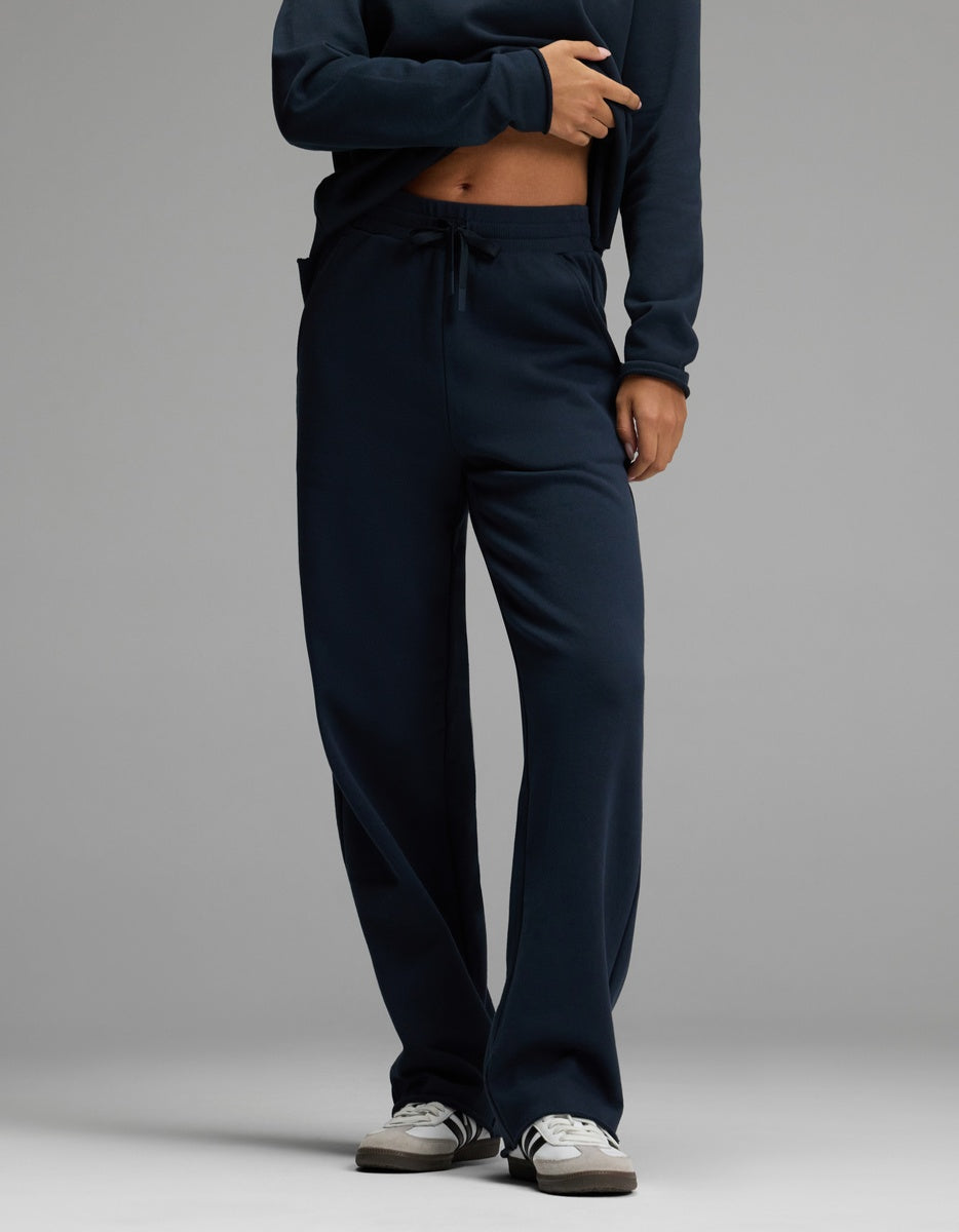 A person wearing Cozy Earth’s Women’s Chelsea Pant in Eclipse, paired with a matching sweatshirt and white sneakers, stands against a plain gray background. Their hand rests near the waistband of the pants, and their face is not visible in the image.|Color:Eclipse