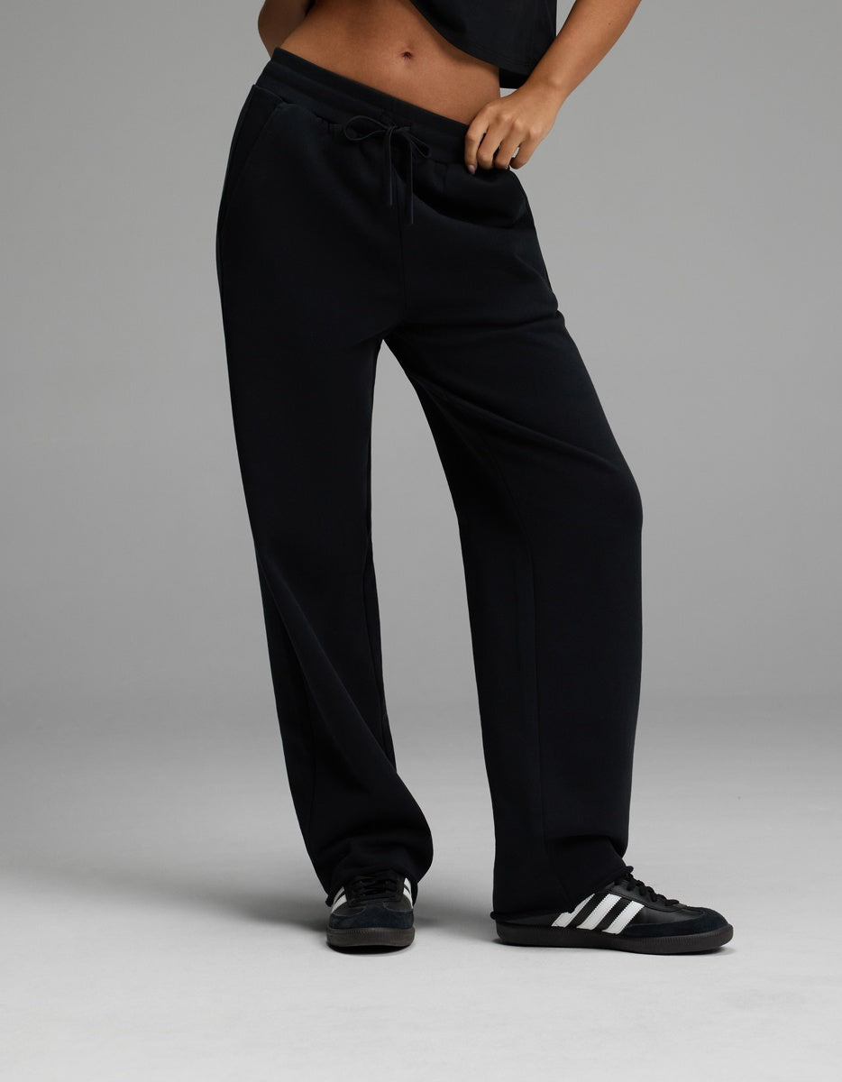 A person wearing the Cozy Earth Women’s Chelsea Pant in Jet Black pairs it with a black top and black sneakers with white stripes, standing with one hand in a pocket against a plain gray background.|Color:Jet Black