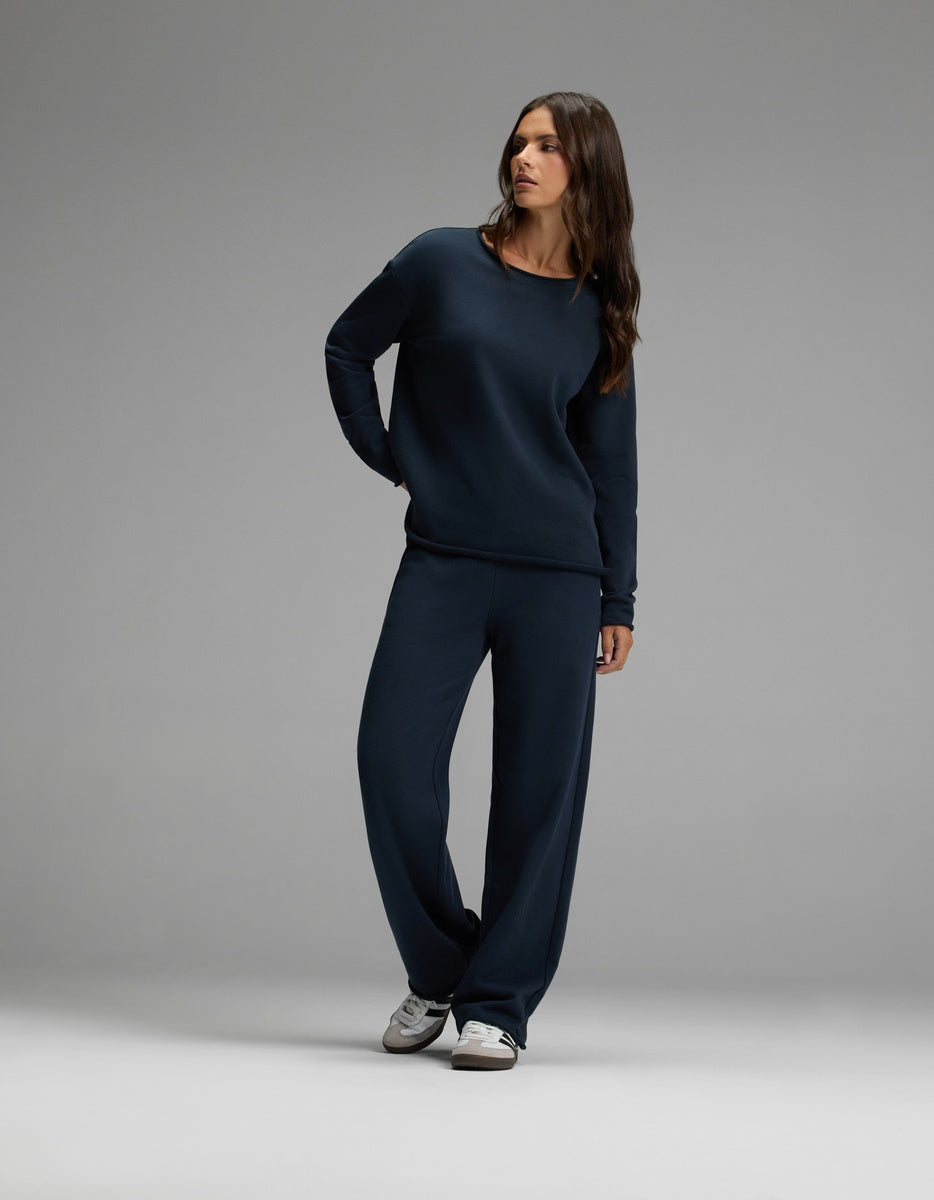 A woman wearing the Cozy Earth Women’s Chelsea Set in Eclipse—featuring a dark blue long-sleeve top and matching wide-leg pants—stands against a plain gray background, looking to the side. She has long brown hair and is wearing white sneakers.|Color:Eclipse