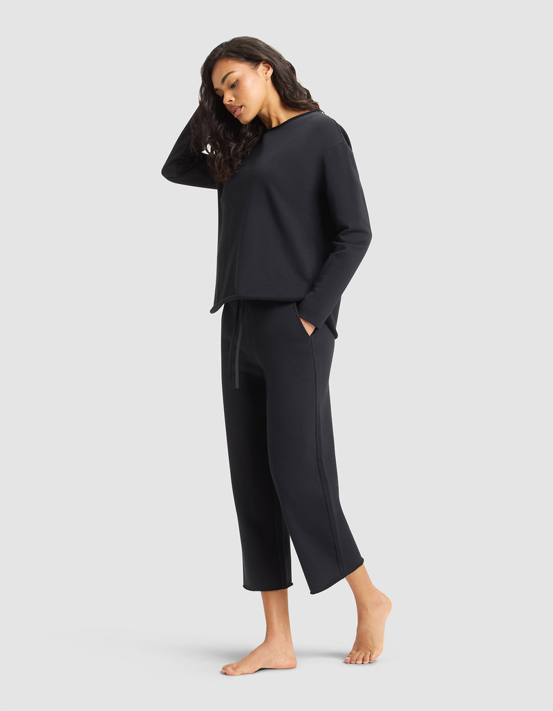A woman with long dark hair stands barefoot against a light gray background, looking relaxed in the Cozy Earth Women's Chelsea Set—wearing a loose-fitting black top and matching cropped pants with her hands in her pockets.