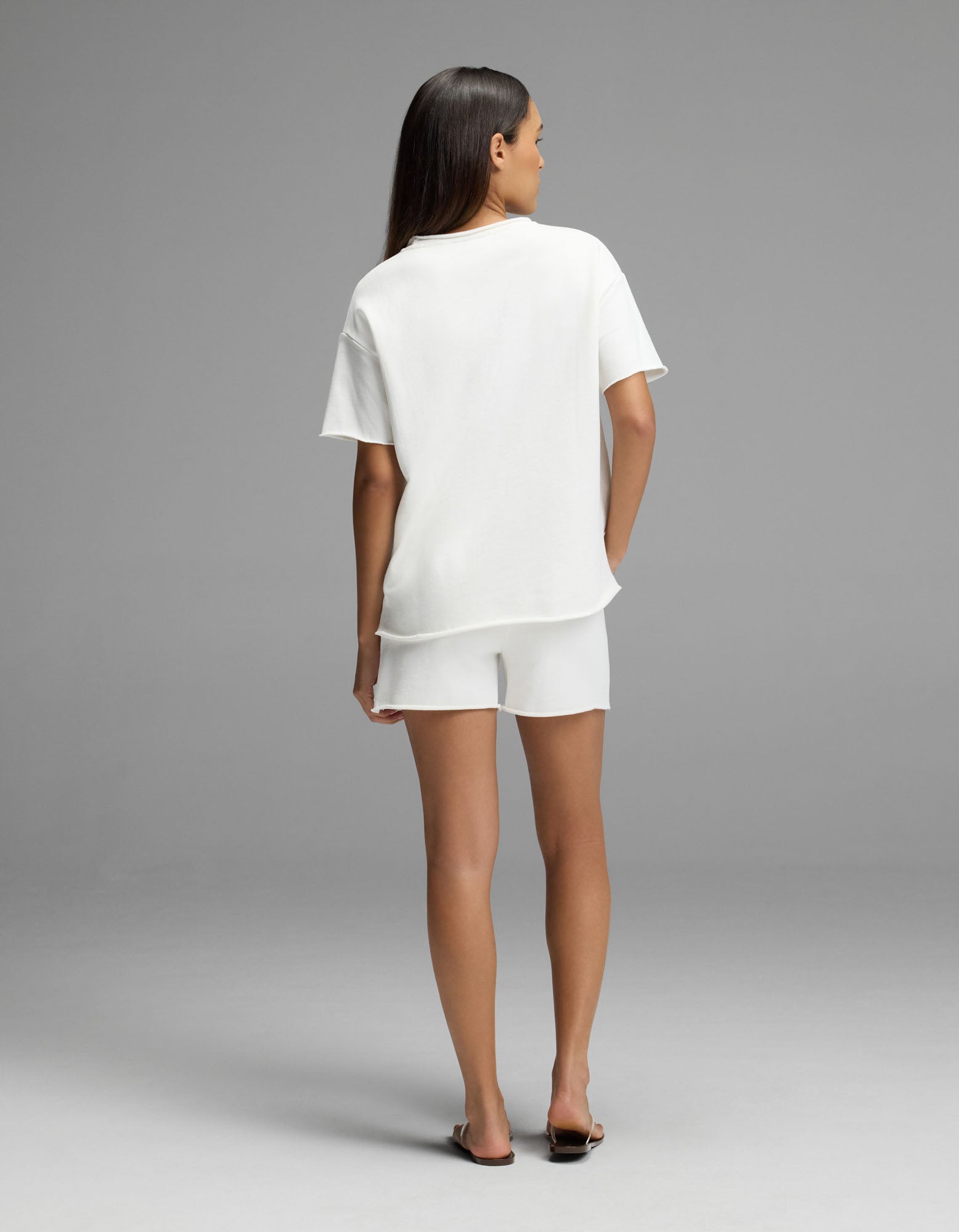 A woman stands facing away in the Cozy Earth Women’s Chelsea Short Sleeve Set, featuring a loose white t-shirt and matching white shorts, paired with black sandals against a plain gray background.|Color:Bone