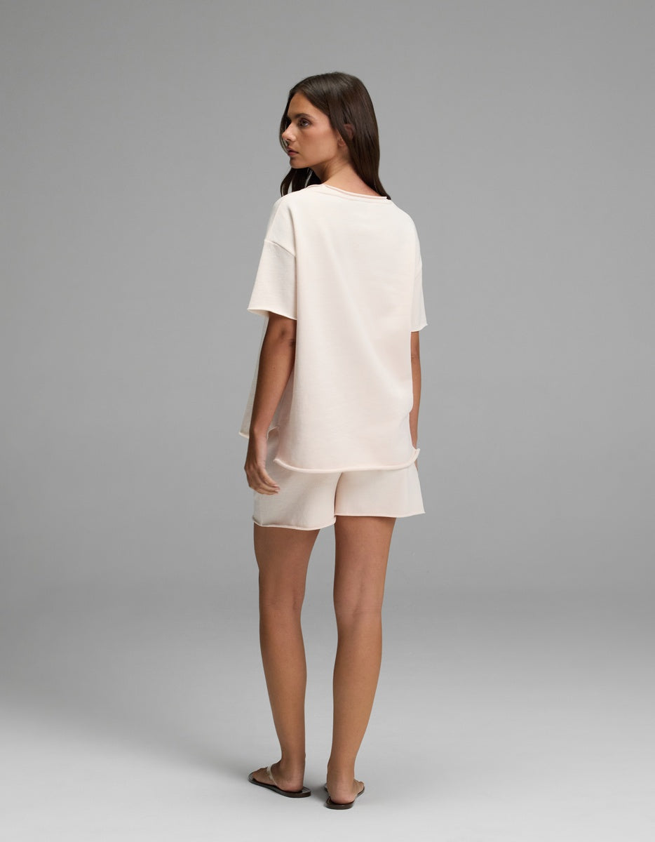 A woman stands facing away, slightly looking over her shoulder, wearing the Cozy Earth Women’s Chelsea Short Sleeve Set in a light color. The background is plain gray.