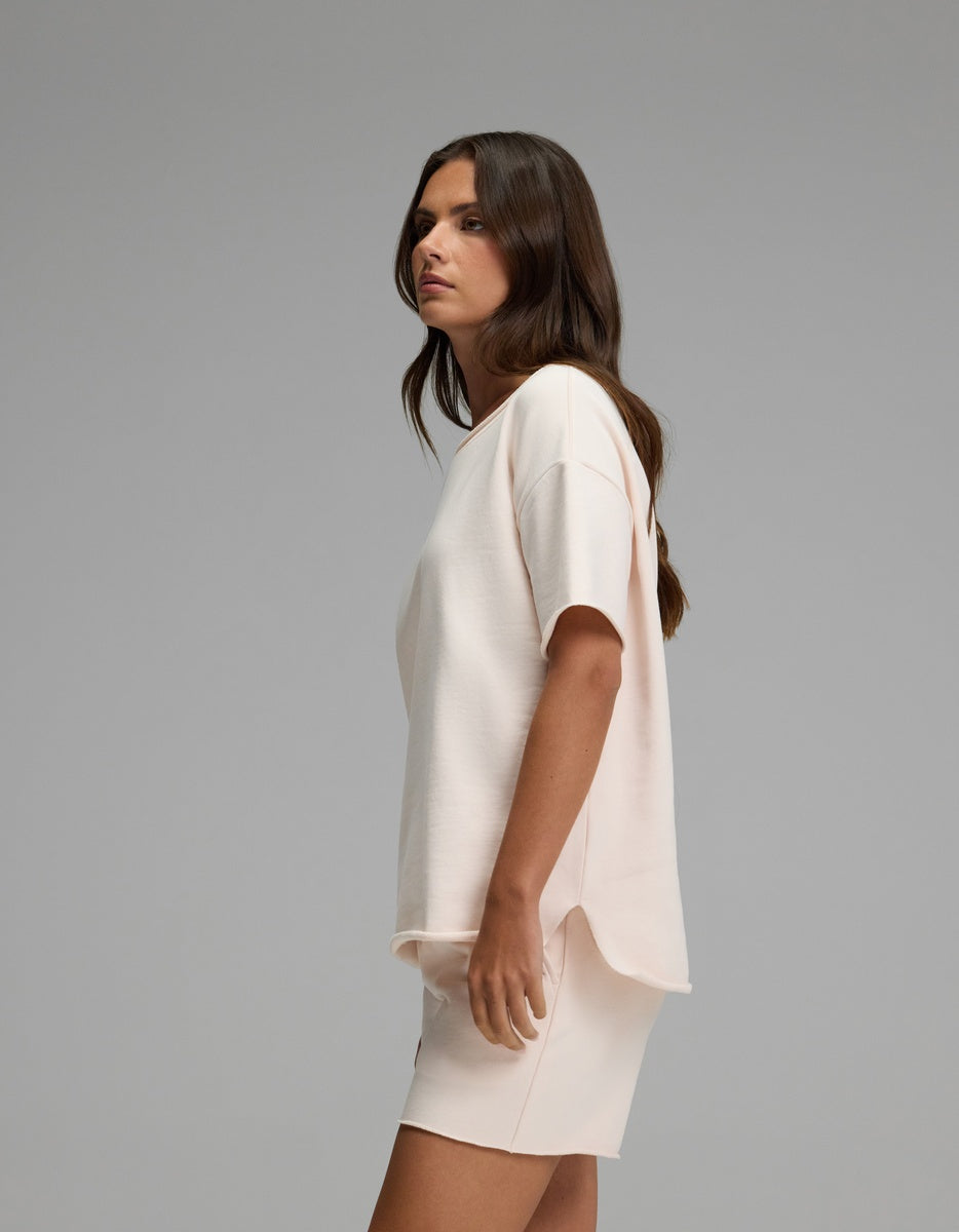 A woman with long brown hair stands sideways against a plain gray background, wearing the Women’s Chelsea Short Sleeve Set in light beige from Cozy Earth.