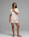 A woman stands against a plain background, barefoot, with long brown hair and one hand touching her collar. She is wearing the Cozy Earth Women’s Chelsea Short Sleeve Set in Petal.|Color:Petal