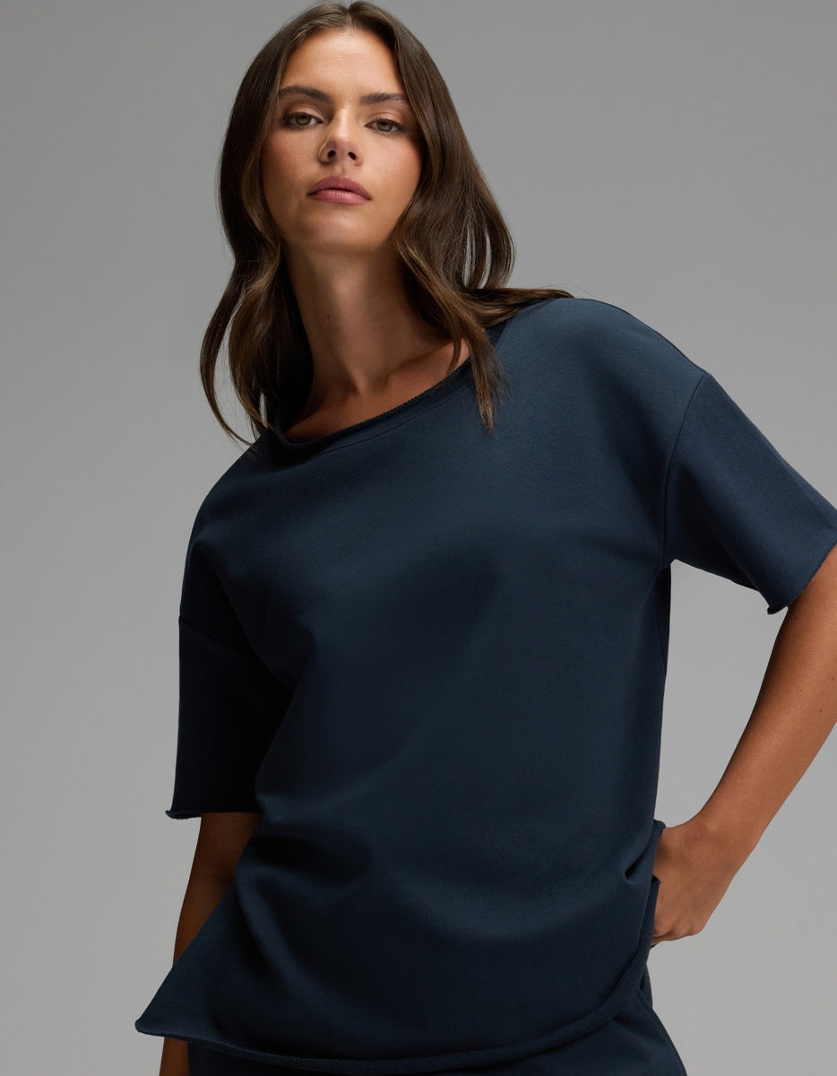 A woman with long brown hair wears the Cozy Earth Women’s Chelsea Short Sleeve in Eclipse, a loose-fitting navy blue top, and poses with one hand in her pocket against a plain gray background.|Color:Eclipse