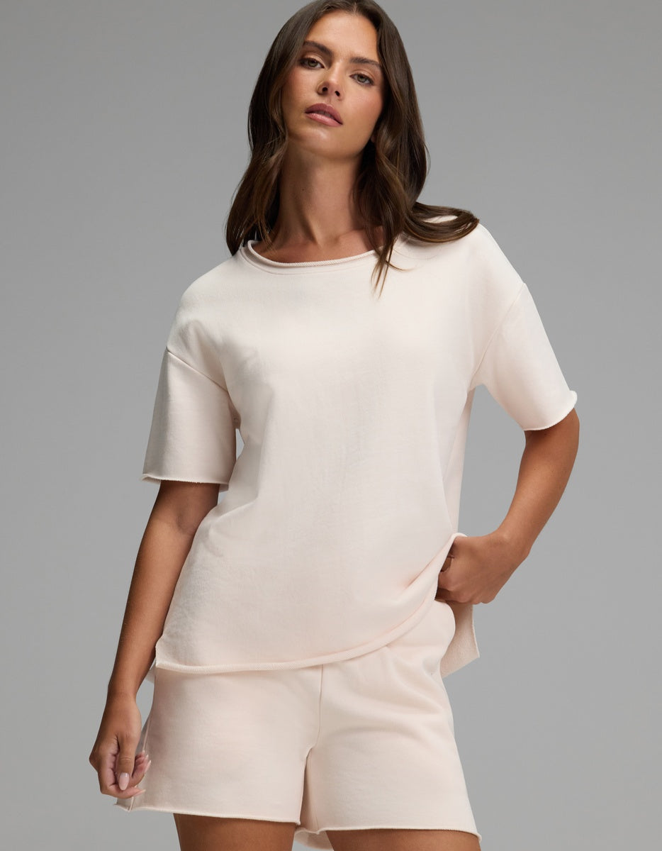 A woman with long brown hair wears the Cozy Earth Women’s Chelsea Short Sleeve in Petal with matching shorts, standing against a plain gray background with one hand in her pocket.|Color:Petal