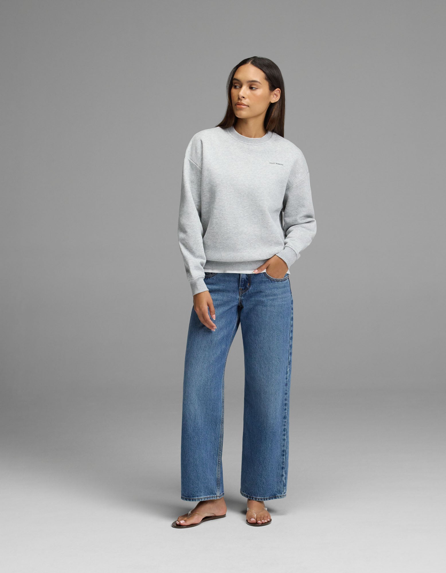 A woman stands against a gray background wearing the Cozy Earth Women’s Cityscape Crewneck, styled with blue wide-leg jeans and beige sandals. Her left hand is in her pocket as she looks slightly to the side.