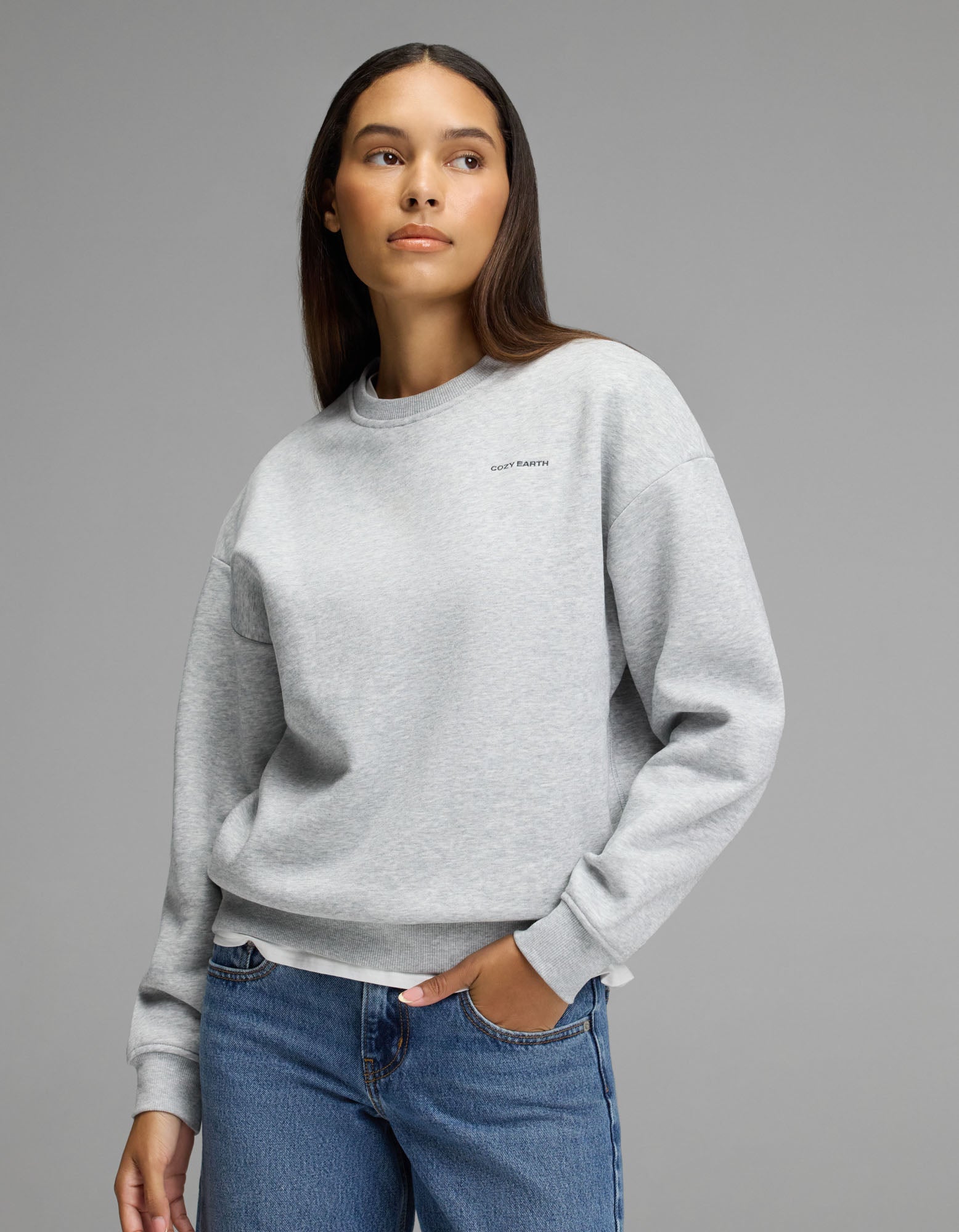 A person models the Cozy Earth Women’s Cityscape Crewneck in Heather Grey, paired with blue jeans, standing against a plain gray background and glancing to the side with one hand in their pocket.|Color:Heather Grey