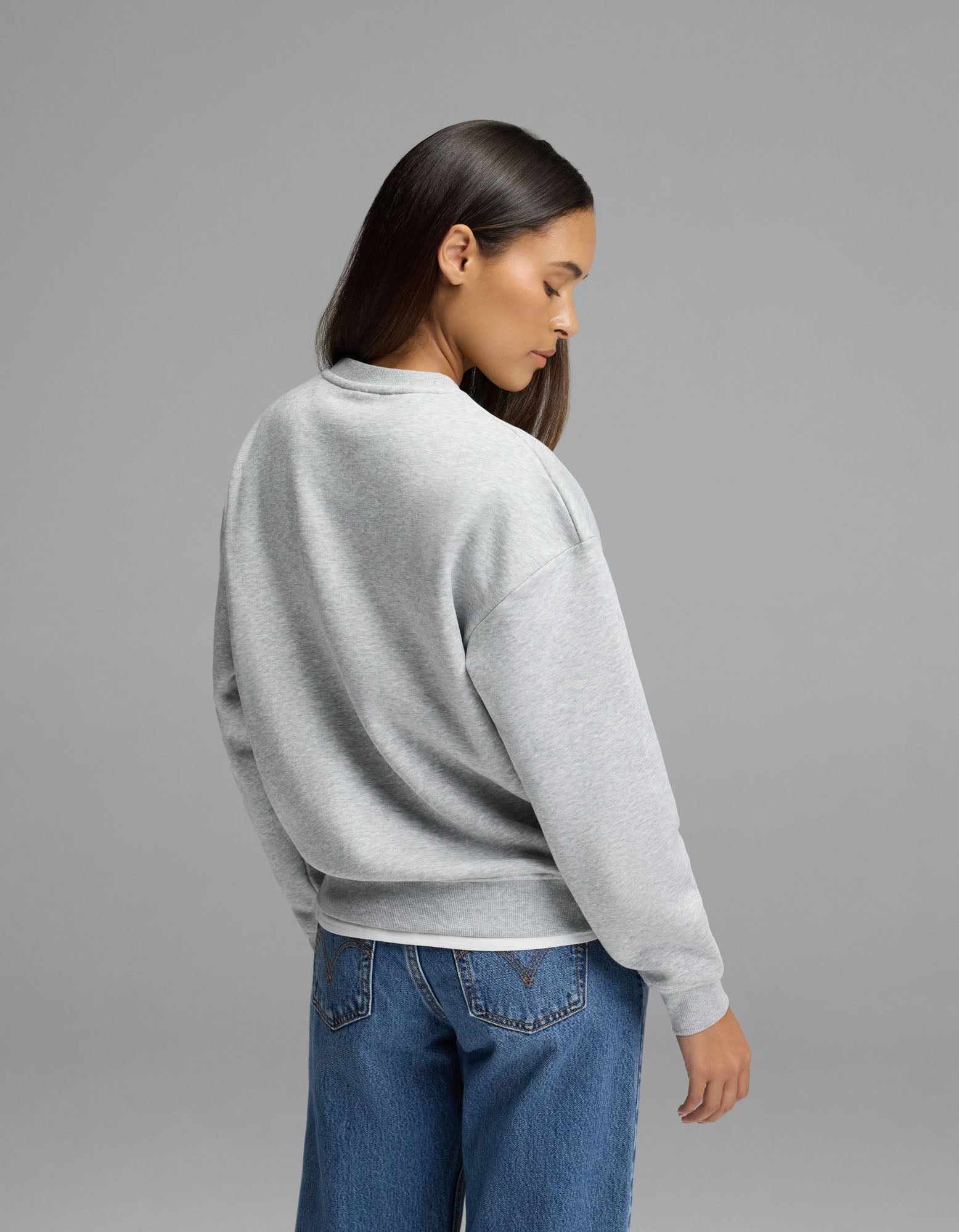 A woman with long dark hair, wearing the Cozy Earth Women’s Cityscape Crewneck in light gray and blue jeans, stands facing away from the camera against a plain gray background.|Color:Heather Grey