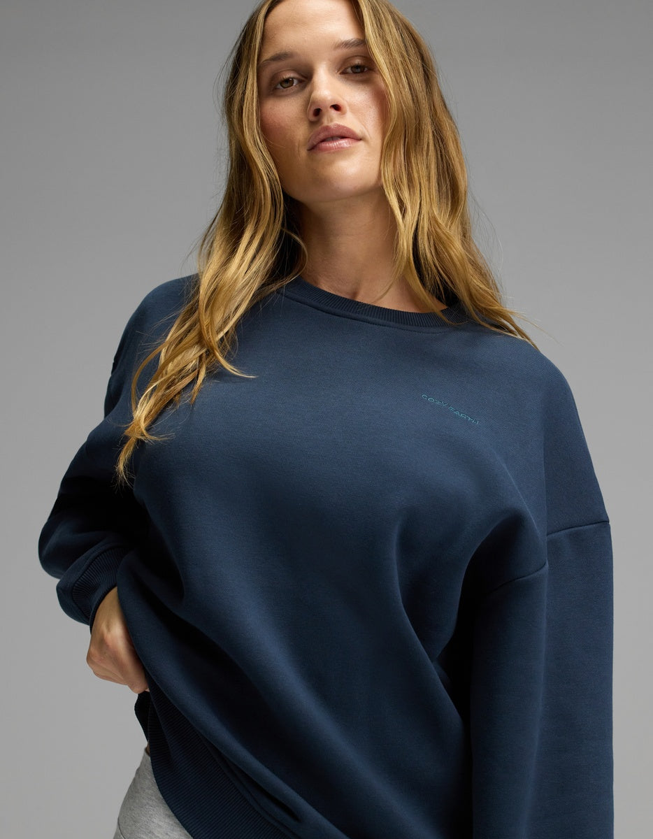 A woman with long, wavy hair stands confidently against a plain gray background, wearing the Cozy Earth Women’s CityScape Crewneck in navy blue paired with light gray pants.|Color:Eclipse