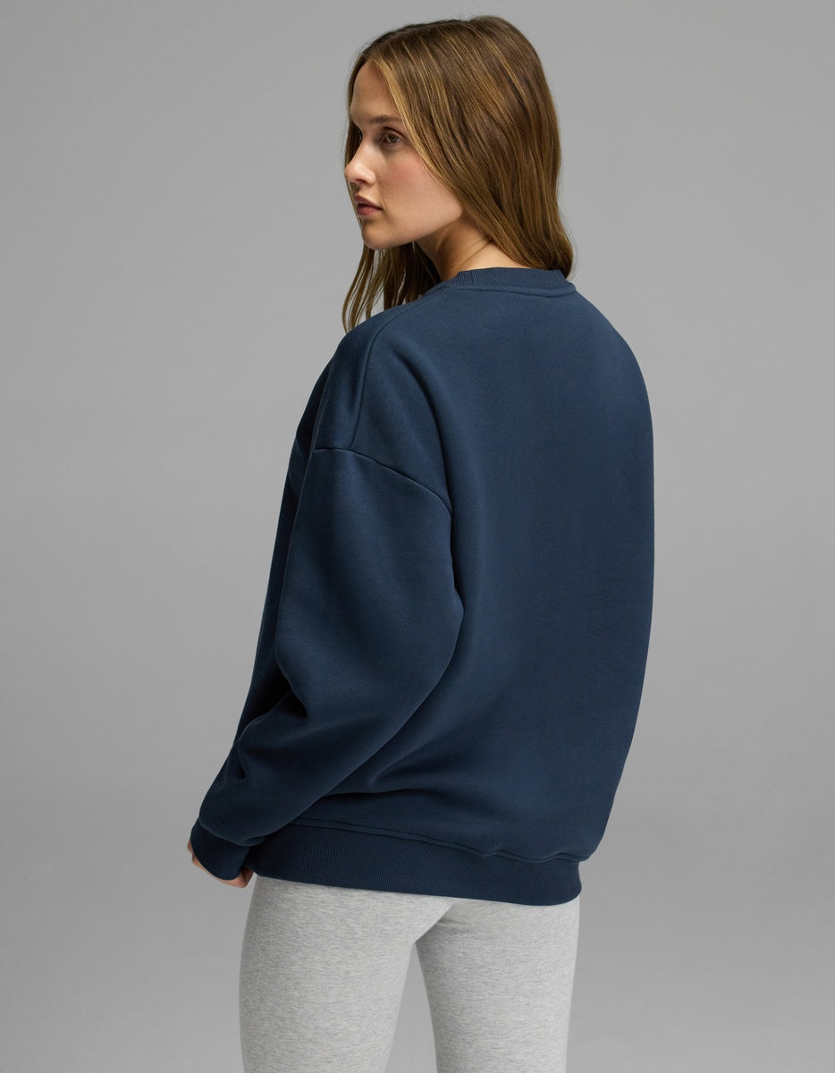 A young woman with long brown hair wearing the Cozy Earth Women’s CityScape Crewneck in navy and light gray leggings stands and looks over her shoulder against a plain gray background.