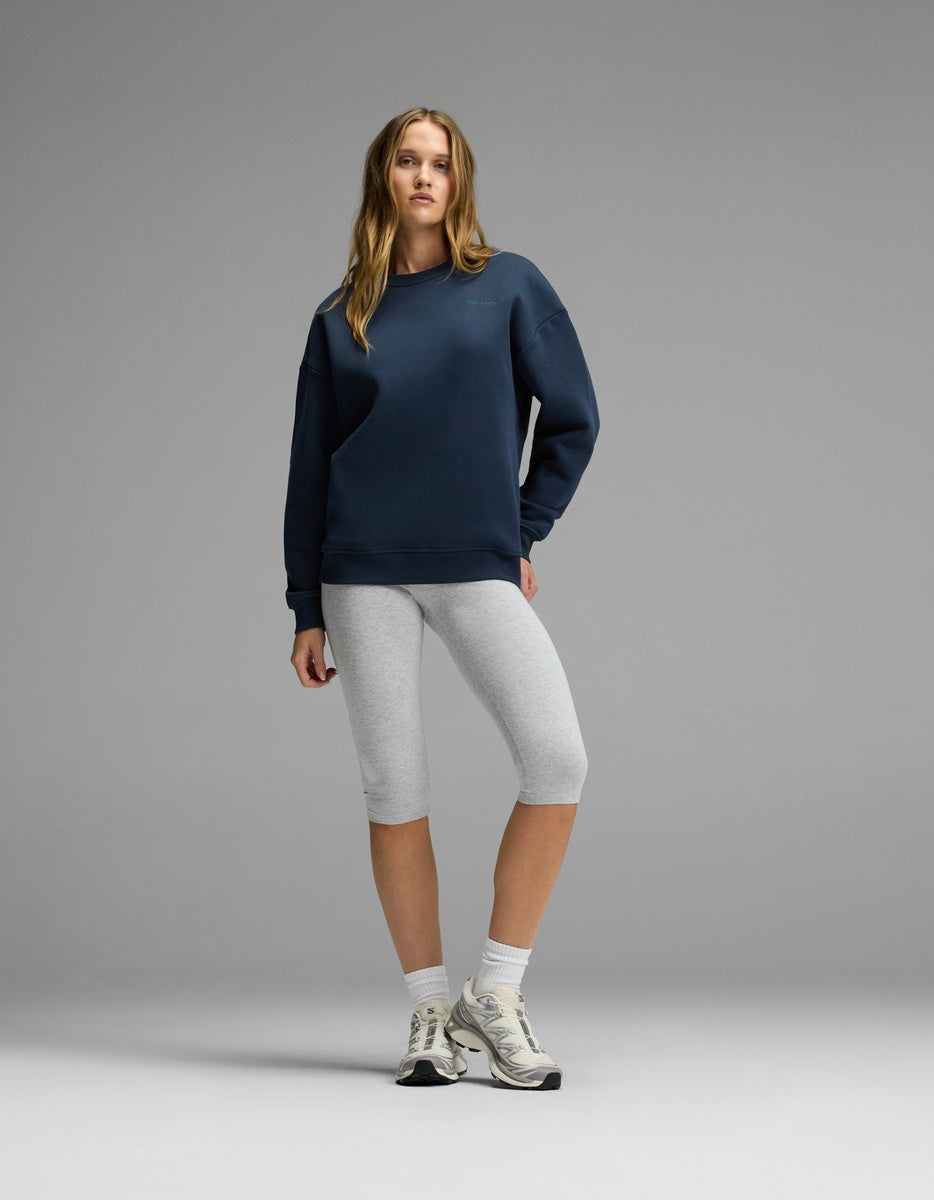 A woman stands against a plain gray background wearing the Cozy Earth Women’s CityScape Crewneck in navy, paired with light gray knee-length leggings, white crew socks, and white sneakers. She has long hair and a relaxed expression, with one hand on her hip.